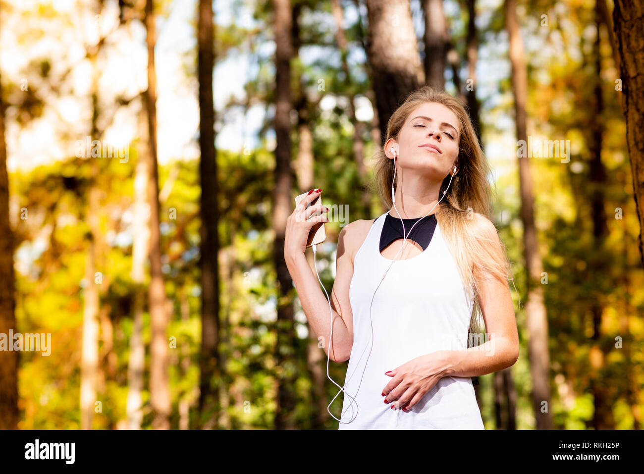 Beautiful blonde model in gym clothes listening to music on her cell phone. Blurred forest background at sunset Stock Photo