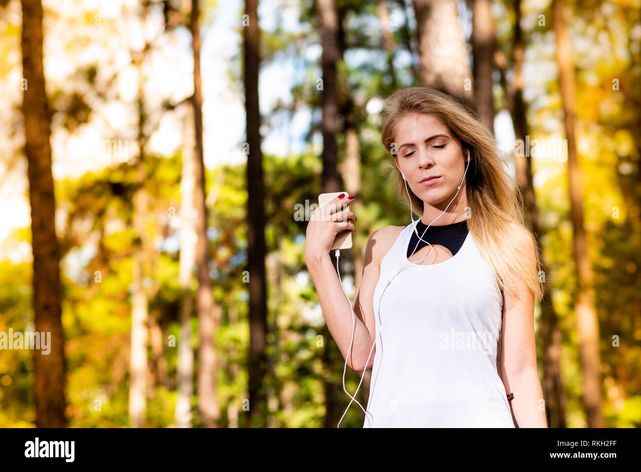 Beautiful blonde model in gym clothes listening to music on her cell phone. Blurred forest background at sunset Stock Photo