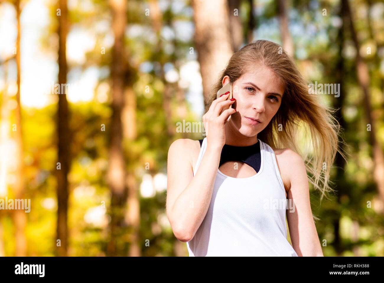 Beautiful blonde model in gym clothes talking on cellphone. Blurred forest background at sunset Stock Photo