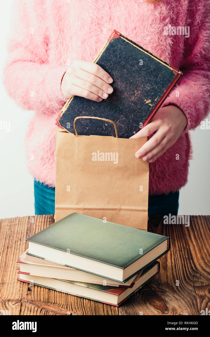 Young girl putting the books into paper bag in bookstore. A few books on a wooden table. Teenager girl wearing pink sweater and blue jeans. Vertical p Stock Photo