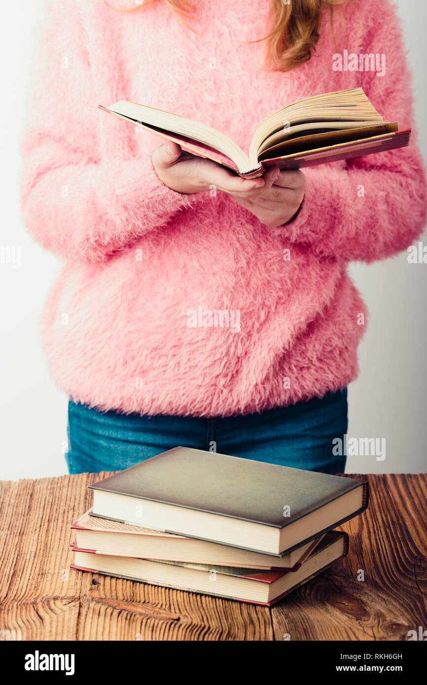 Young girl holding a book standing in bookstore. A few books on a wooden table. Teenager girl wearing pink sweater and blue jeans. Vertical photo Stock Photo