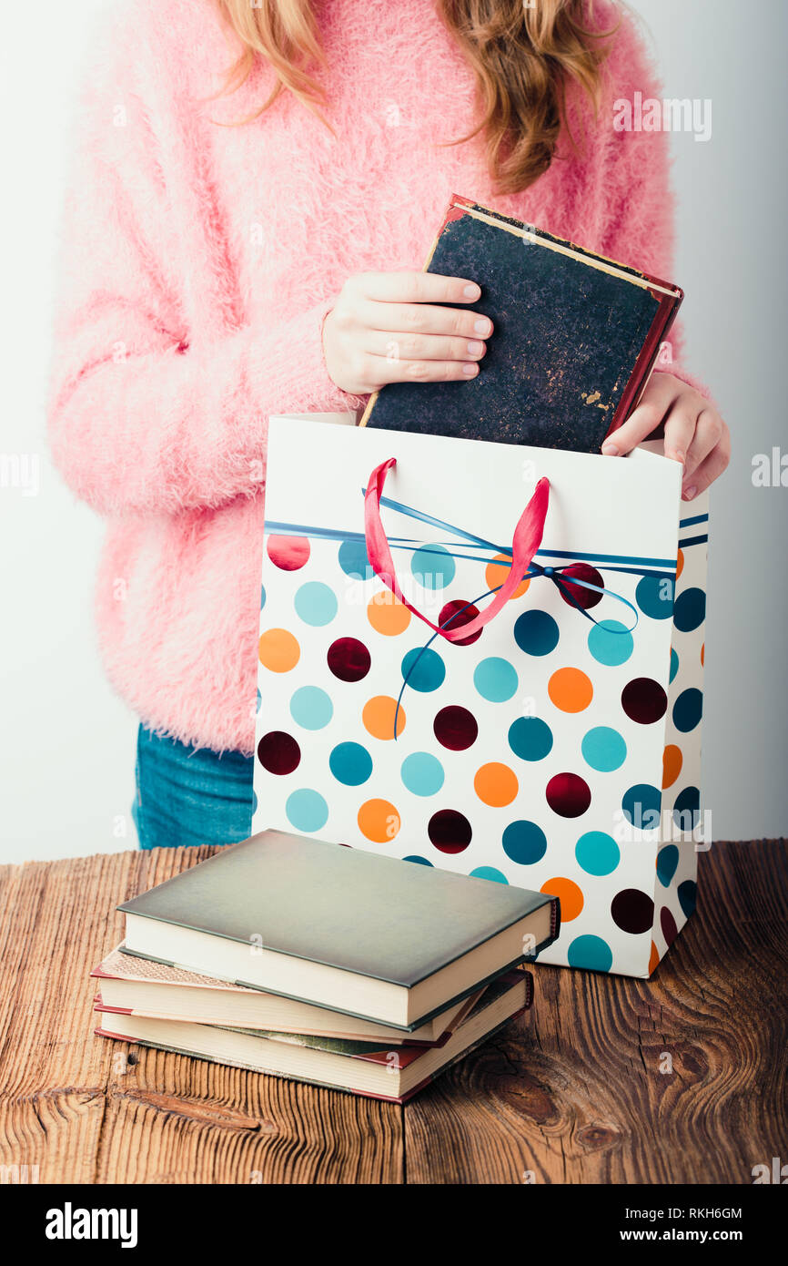 Young girl putting the books into paper bag in bookstore. A few books on a wooden table. Teenager girl wearing pink sweater and blue jeans. Vertical p Stock Photo