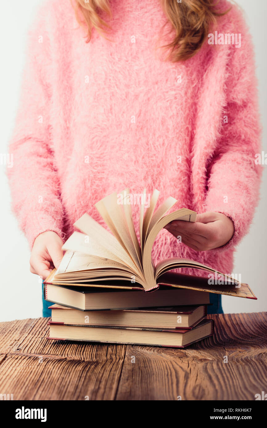 Young girl turning the pages of book in library A few books on a wooden table. Teenager girl wearing pink sweater and blue jeans. Vertical photo Stock Photo