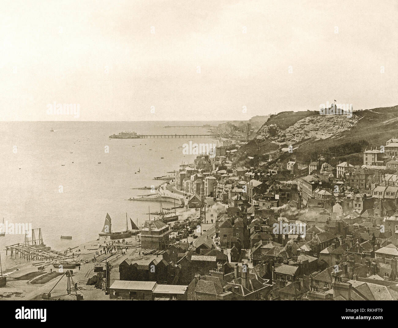 Looking west along the sea front at Hastings, East Sussex, England in the Victorian era, c.1900. In the foreground is the Old Town and both Hastings and St Leonards piers are visible in the background. The shingle beach to the left is The Stade, home to its beach-launched fishing fleet. The distinctive tall, black huts (bottom centre) were used to house the nets for catching the mackerel and herring. Stock Photo