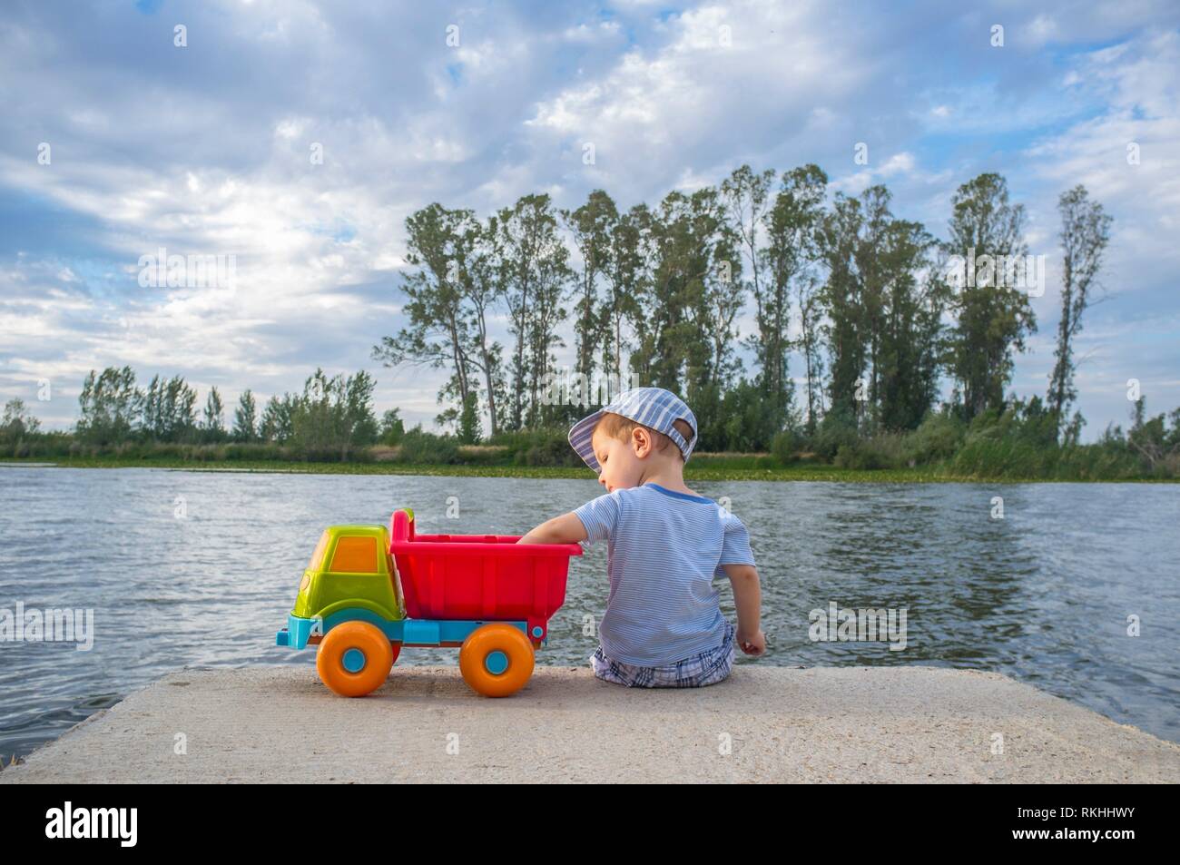 Two year old boy playing at riverside with dump truck. He is sitting over fisher platform throwing stones. Stock Photo