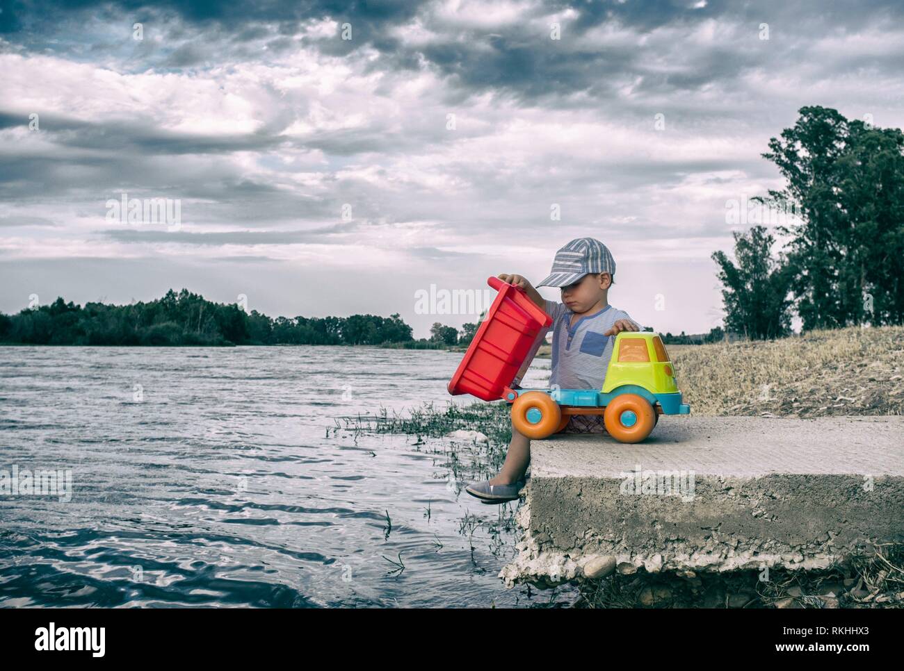 Two year old boy playing at riverside with dump truck. He is unloading stones on water. Stock Photo