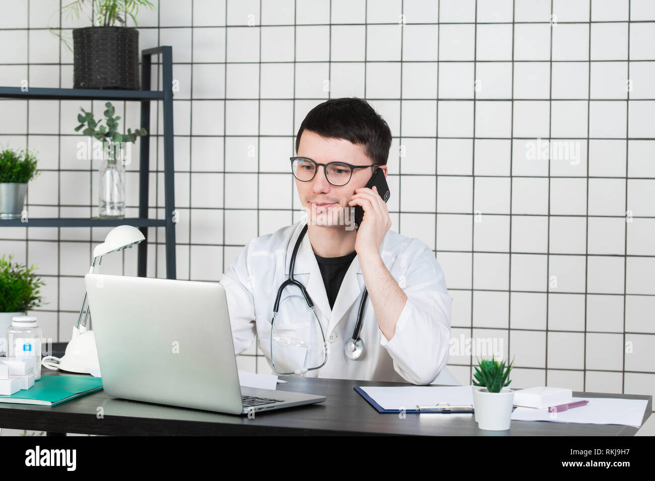 Male doctor using telephone while working on computer at table in clinic Stock Photo