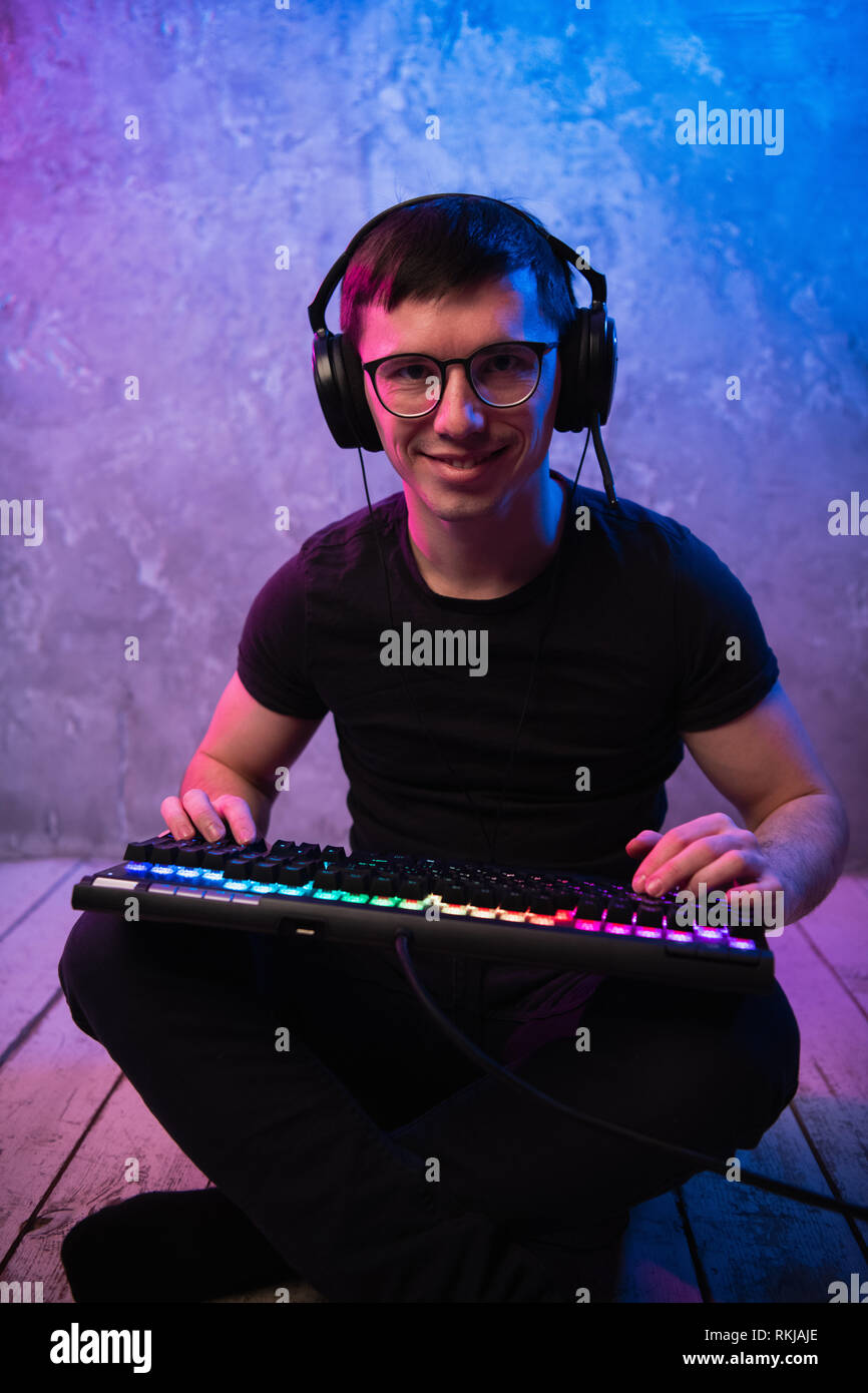 Portrait of the young handsome pro gamer sitting on the floor with keyboard in neon colored room Stock Photo