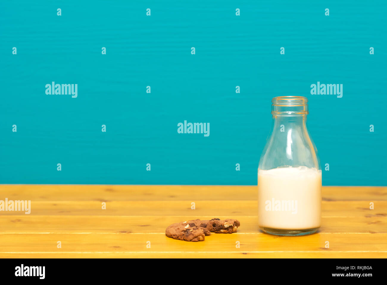 One-third pint glass milk bottle half full with fresh creamy milk and a half-eaten chocolate chip cookie, on a wooden table against a teal background Stock Photo