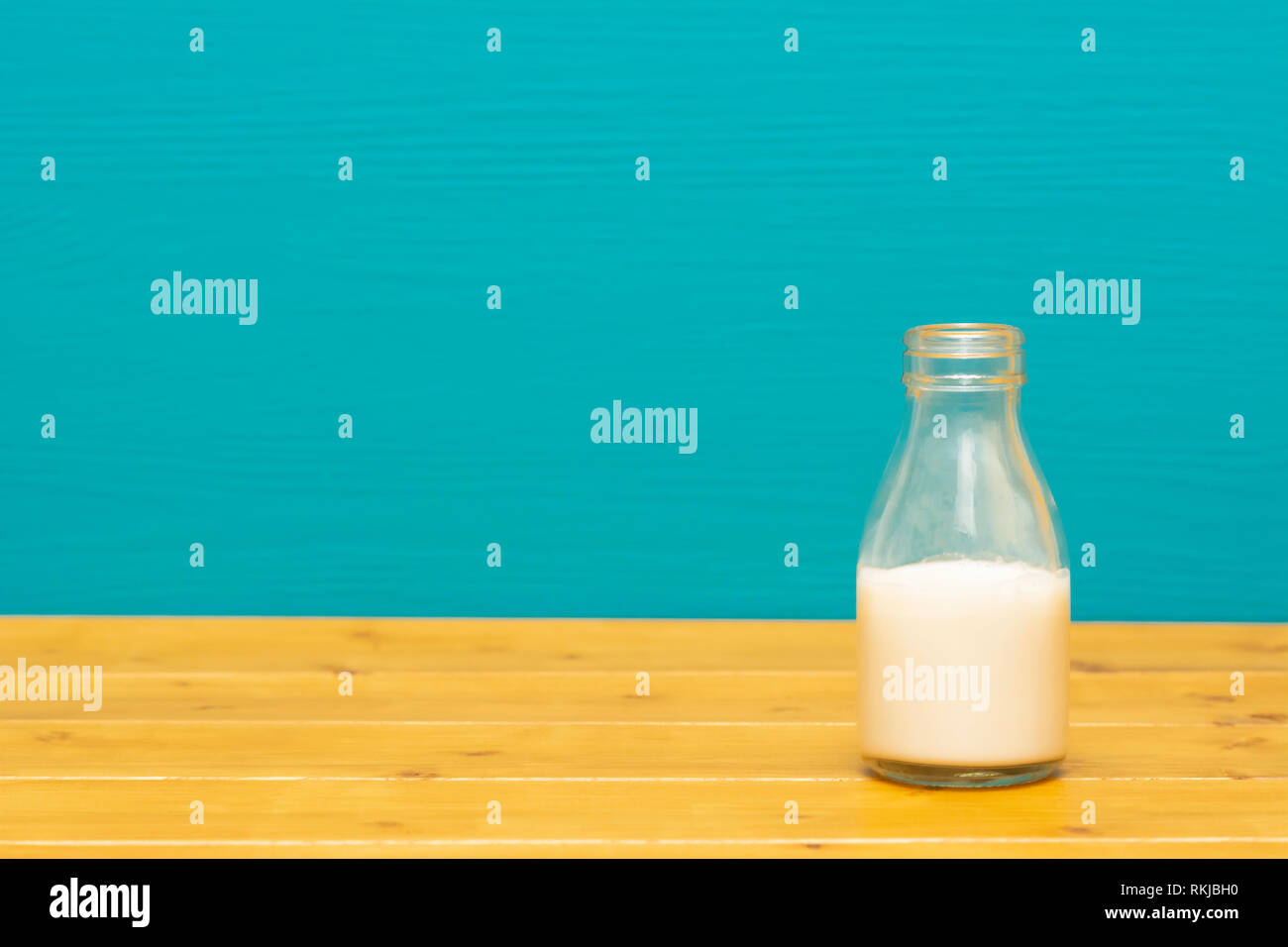 One-third pint glass milk bottle half full with fresh creamy milk, on a wooden table against a bright teal painted background Stock Photo