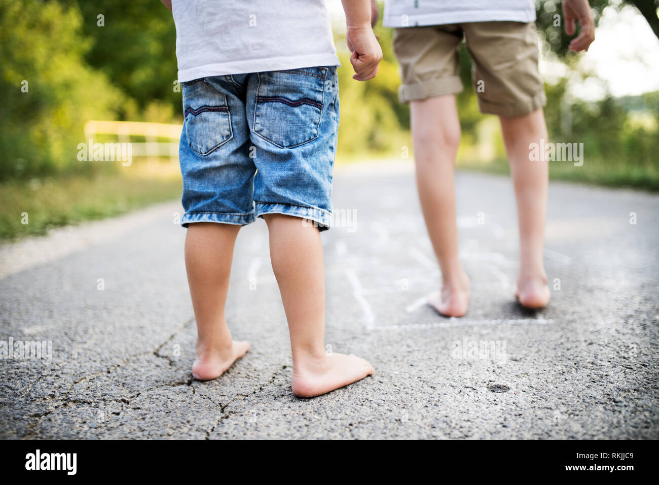 A rear view of two small boys hopscotching on a road in park on a summer day. Stock Photo