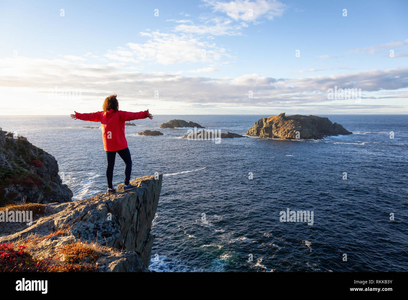 Woman in red jacket is standing at the edge of a cliff with open arms ...