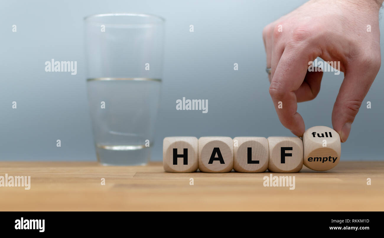 Hand turns a dice and changes the expression 'half empty' to 'half full'. A half full glass of water is standing in front of a grey background. Stock Photo