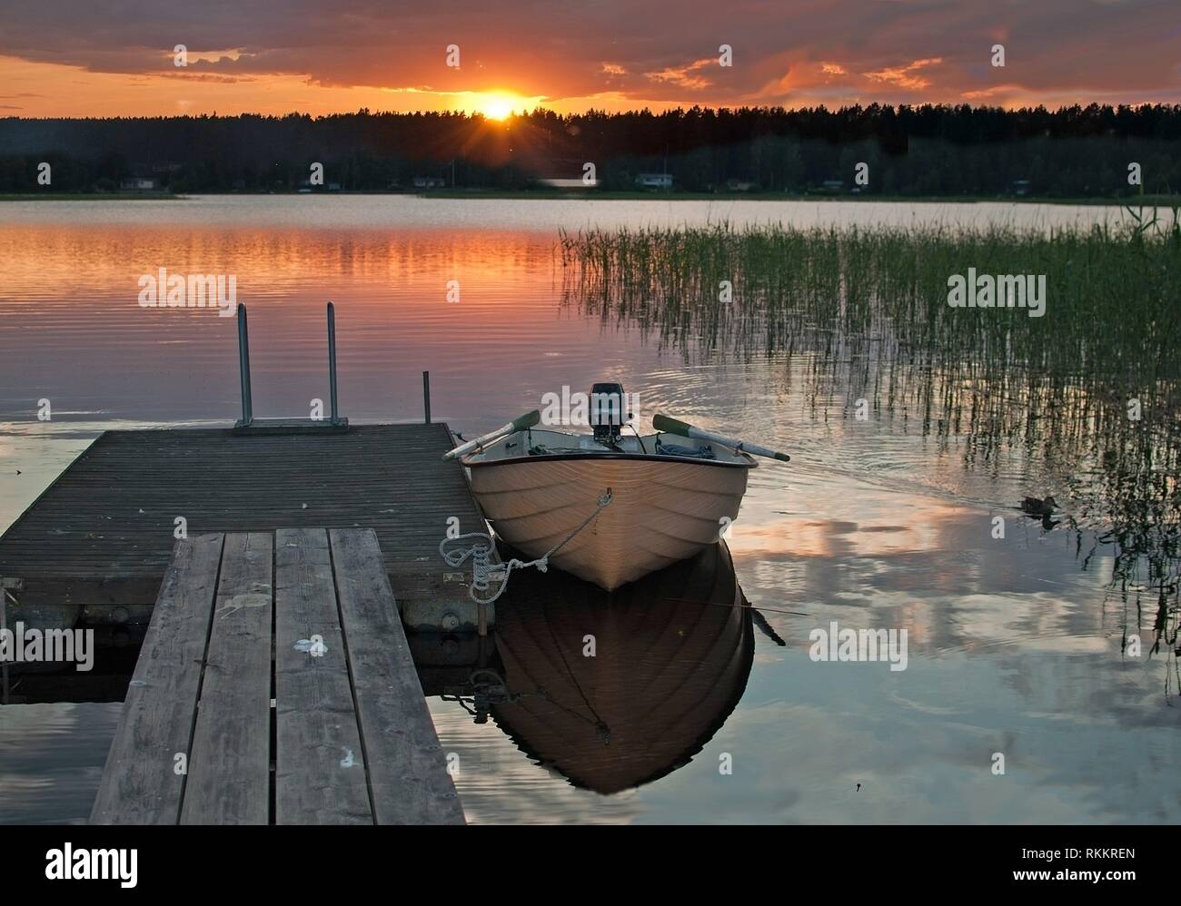 Small boat moored by wooden jetty at peaceful colorful sunset by a lake with sky reflections in tranquil water in Varmland, Sweden. Stock Photo