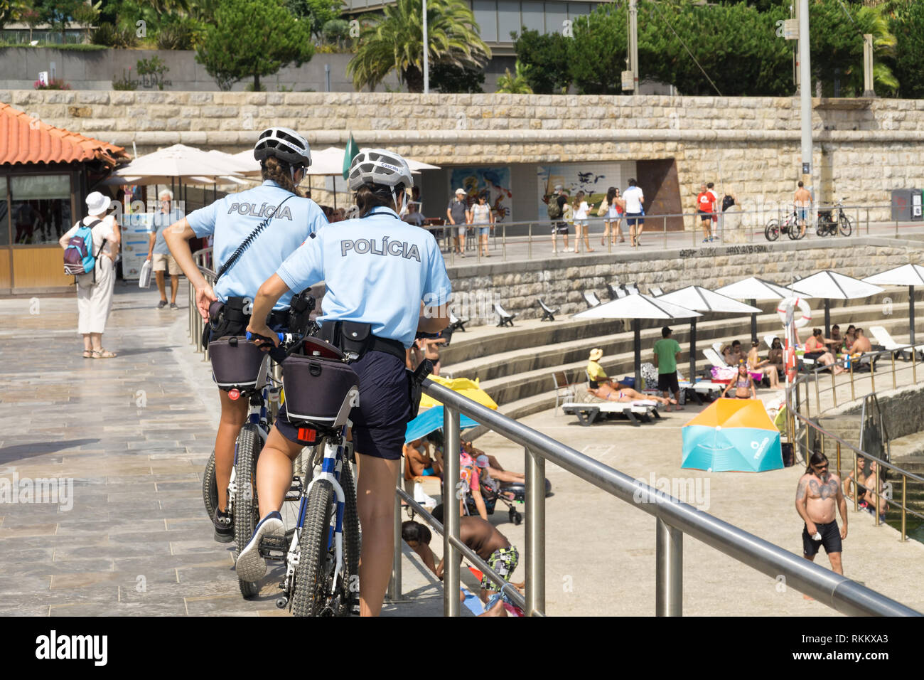 Two policewomen of Cascais, are patrolling seaside promenade  on bicycles. People are sunburning on the city  public beach on the Atlantic shore. Stock Photo