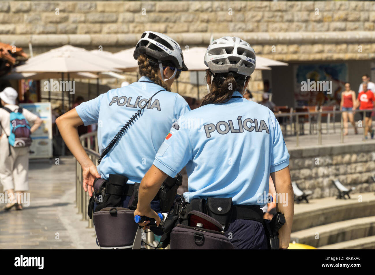 Two policewomen of Cascais, are patrolling seaside promenade  on bicycles. People are sunburning on the city  public beach on the Atlantic shore. Stock Photo