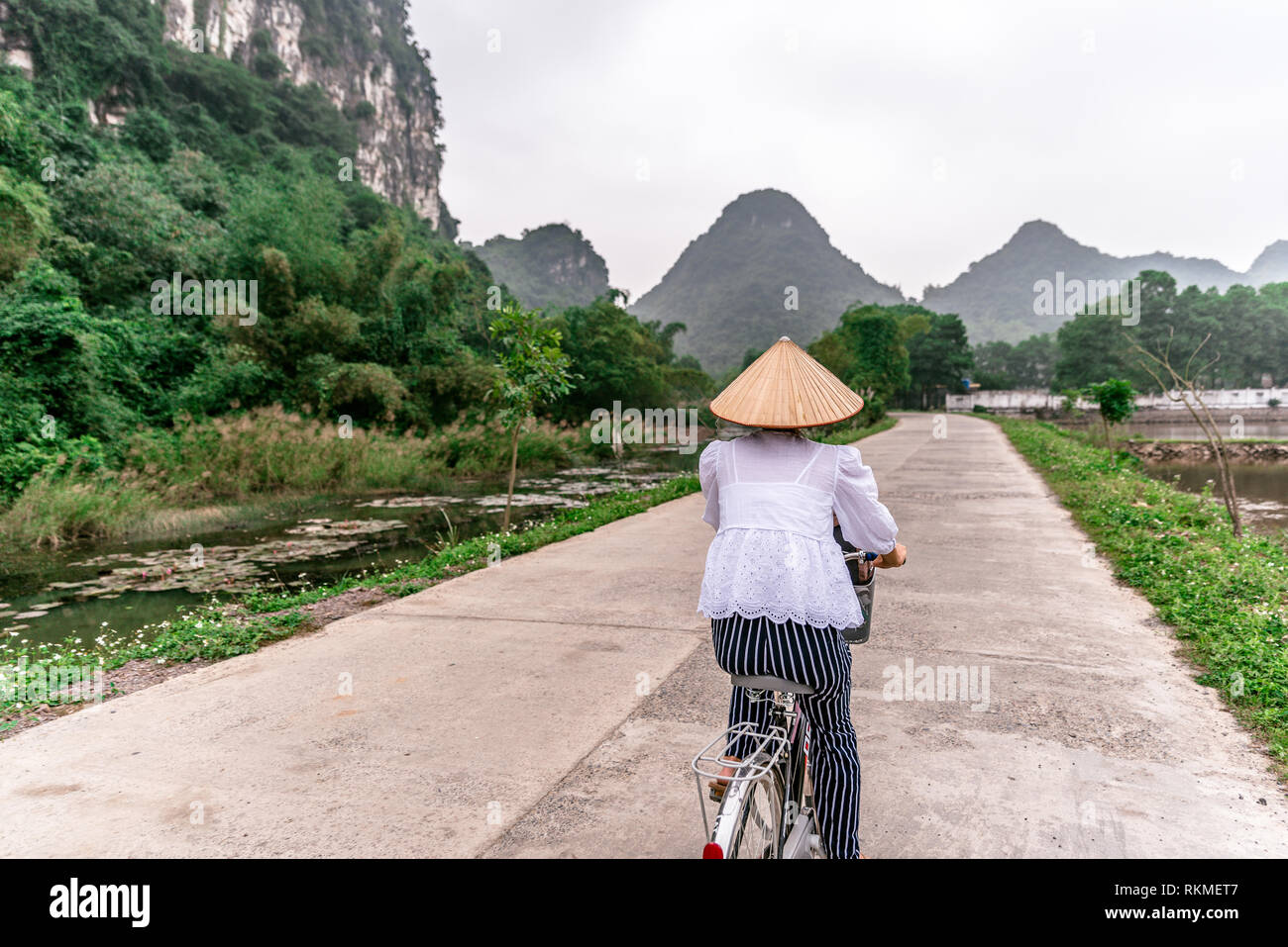 Vietnamese woman with traditional conical straw hat on bicycle. Beautiful Landscape of rice fields and mountain Scenery at Nature reserve Trang An and Stock Photo