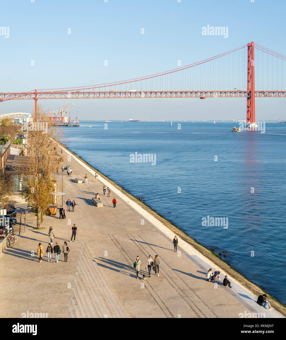 Lisbon, Portugal - 12/28/18: Pedestrian walkway riverside, tagus river. View of the 25 april red bridge, lots of people passing by walking Stock Photo