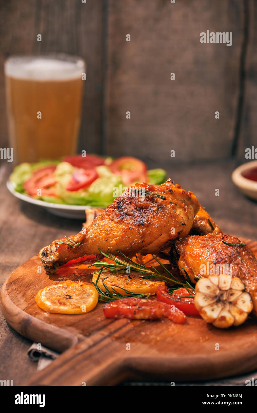 Grilled chicken legs roasted on the grill on wooden chopping board with tomato sauce in a bowl, fresh tomatoes and lettuce leaves, bitter pepper, glas Stock Photo