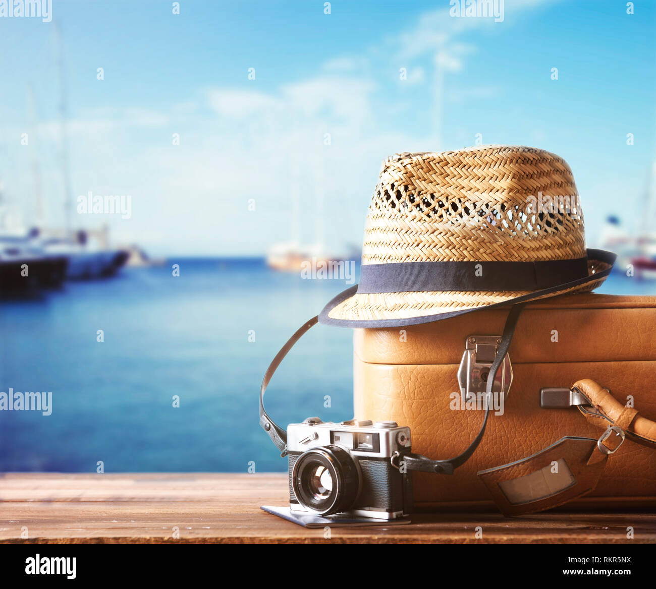 Vintage suitcase, hipster hat, photo camera and passport on wooden dack. Tropical sea, beach and yachts in background. Summer holiday  and cruise trav Stock Photo