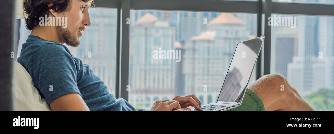 Young man is working on a laptop in his bed on a background of a panoramic window overlooking the skyscrapers. Freelancer, remote work, work from home Stock Photo