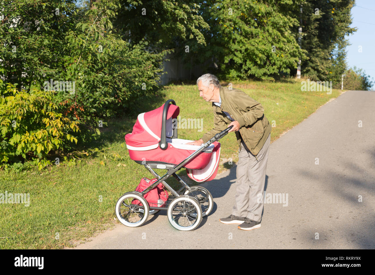 Grandfather carries granddaughter in baby carriager at sunset. Grandfather leaned over to the baby. Outdoor in the country. Stock Photo