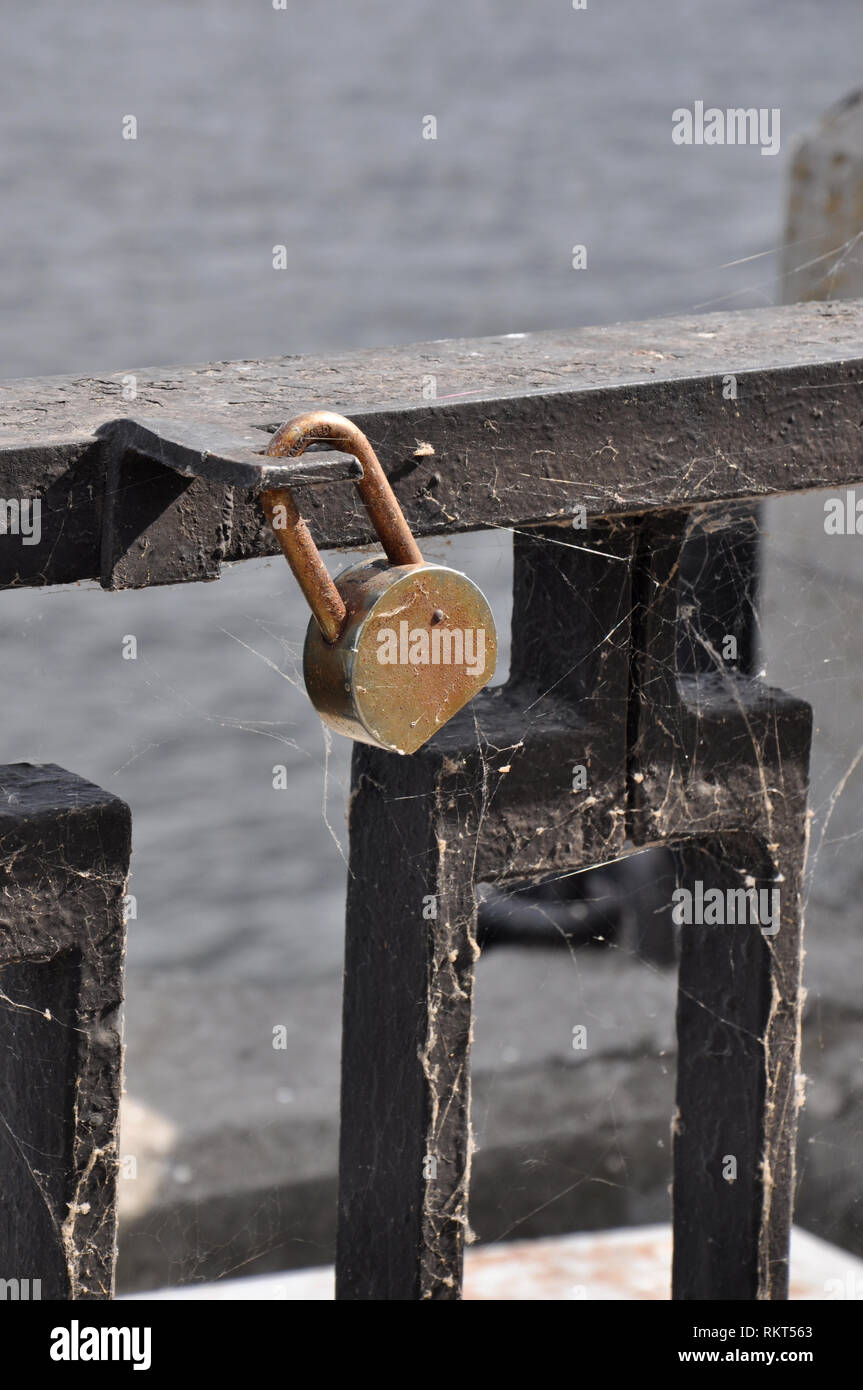 Old rusty padlock hanging on a black bar of an iron parapet covered with cobweb at a river port. Stock Photo