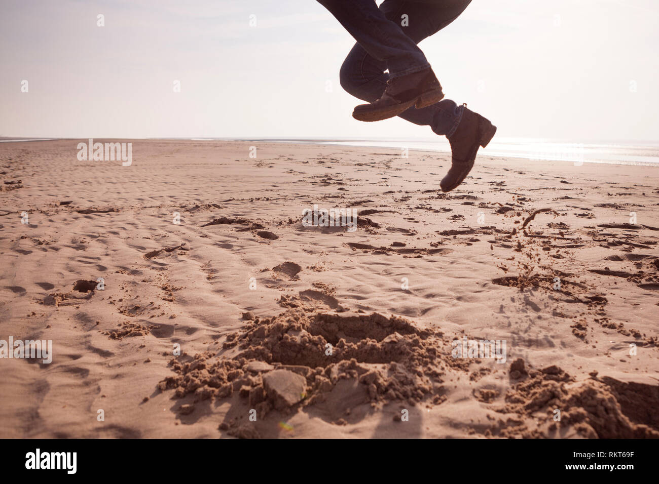 Man wearing jeans and heeled boots, jumping up on a sandy UK beach on a cold spring day Stock Photo