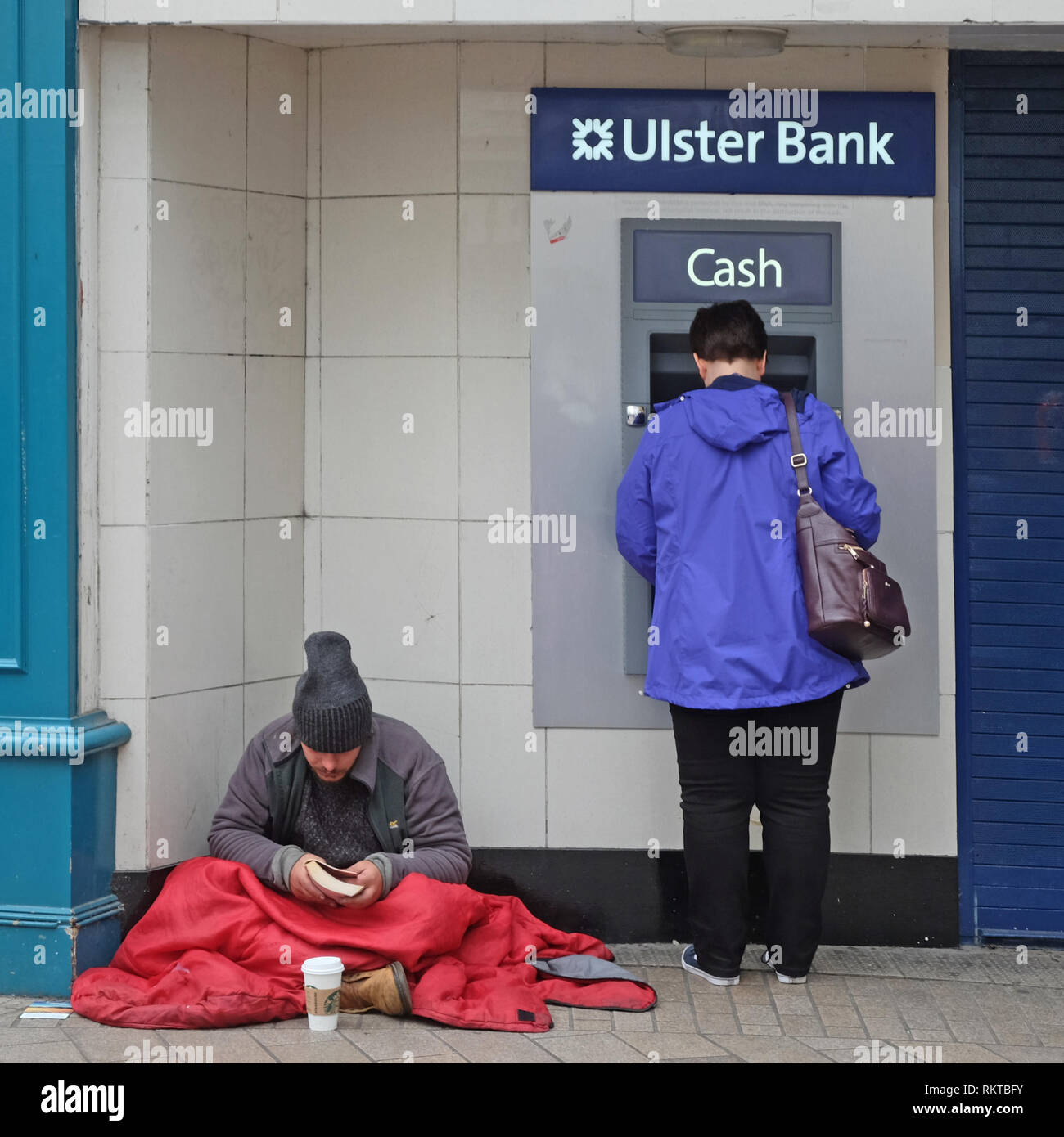 Homeless man reading by Ulster Bank cash point. Stock Photo