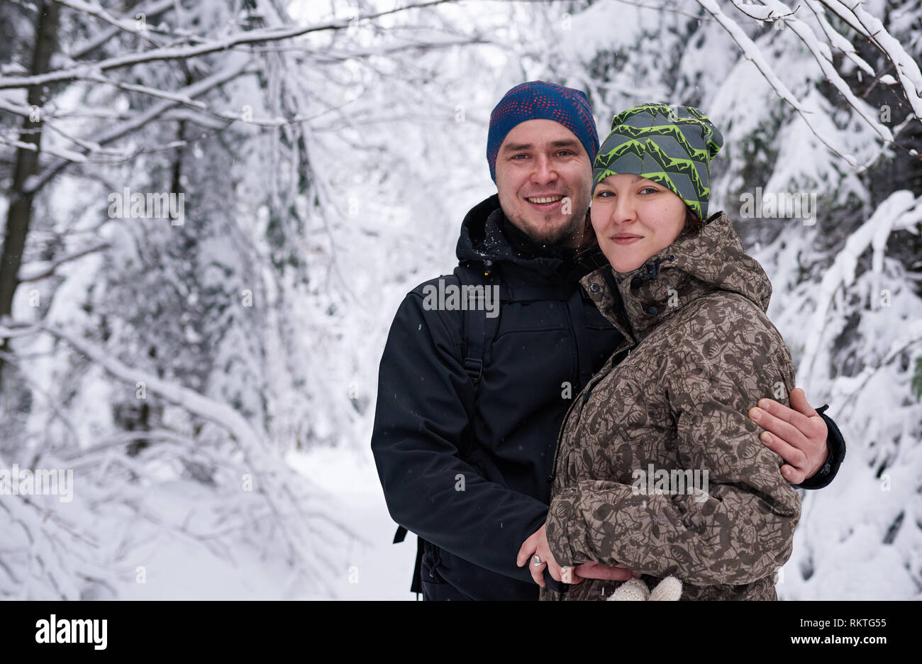 Portrait of a smiling young couple in hiking gear standing arm in arm together in a snow covered forest while out for a winter hike Stock Photo
