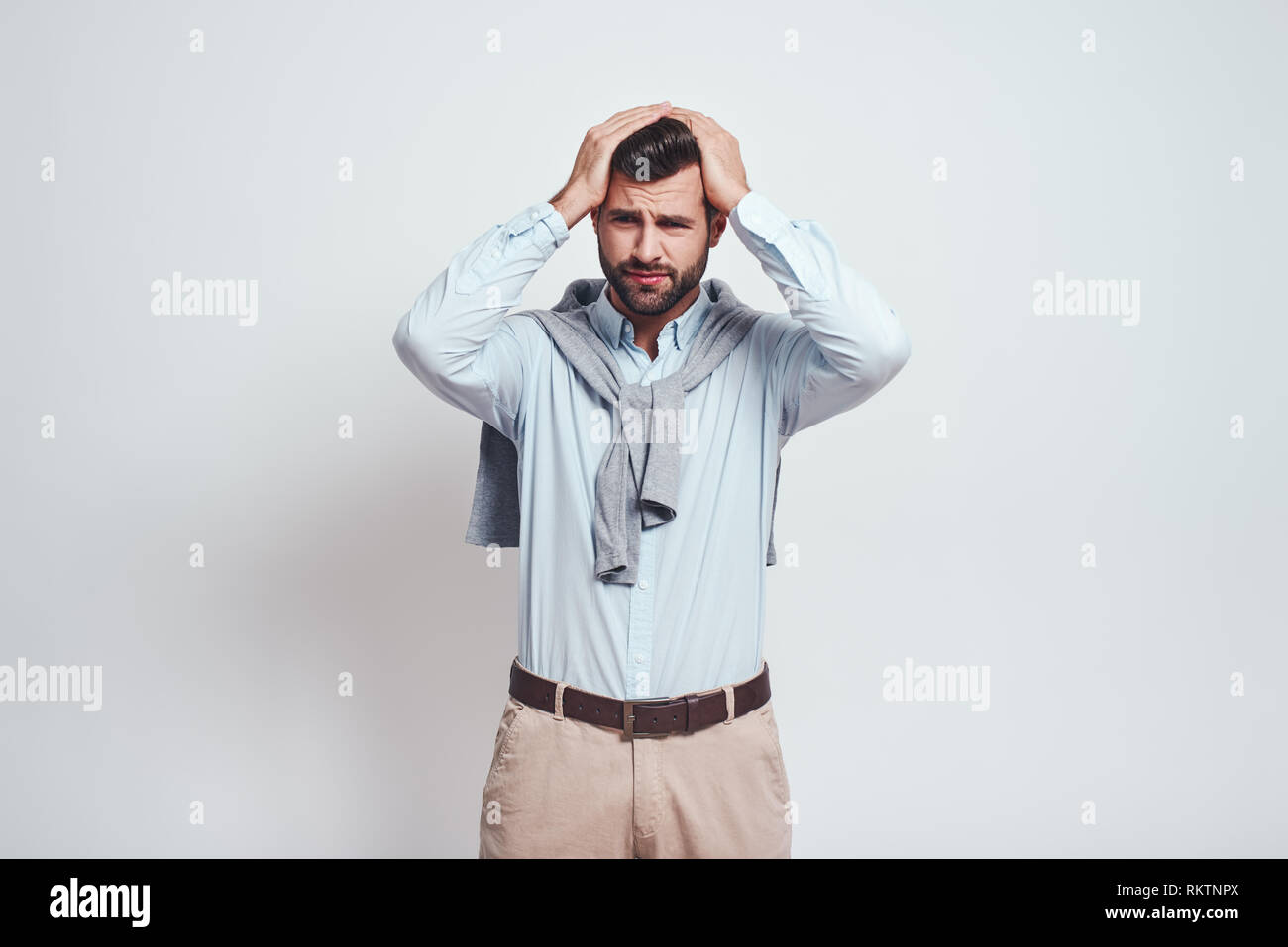Emotional stress. Close-up portrait of confused young man keeping head in his hands and making a sad emotion while standing against grey background. Stress concept. Emotion expression. Stock Photo
