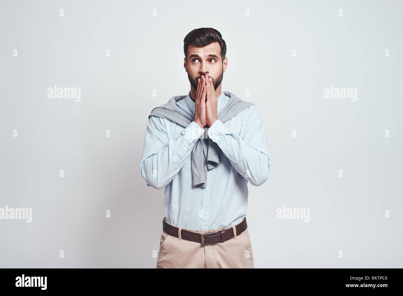 Oops. Confused young bearded man is covering mouth with hands while standing against grey background. Close-up portrait. Emotion expression. Stock Photo
