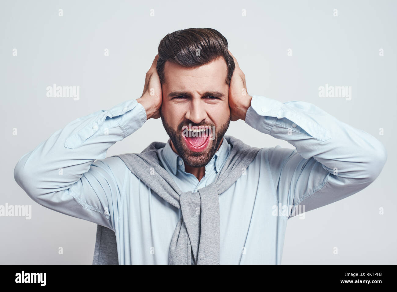 Bla bla bla. Young handsome man is stressed by noise, closing ears with both hands while standing on a grey background. Stress emotion. Stock Photo