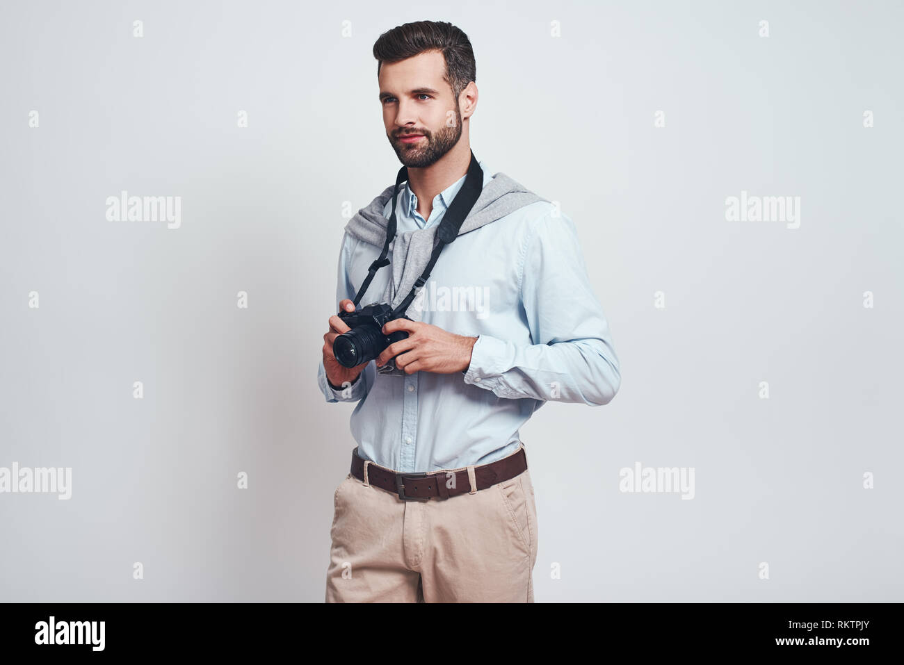 Creative man. Charming young man is holding digital camera and looking away while standing in studio on a grey background. Close-up. Hobby concept. Stock Photo