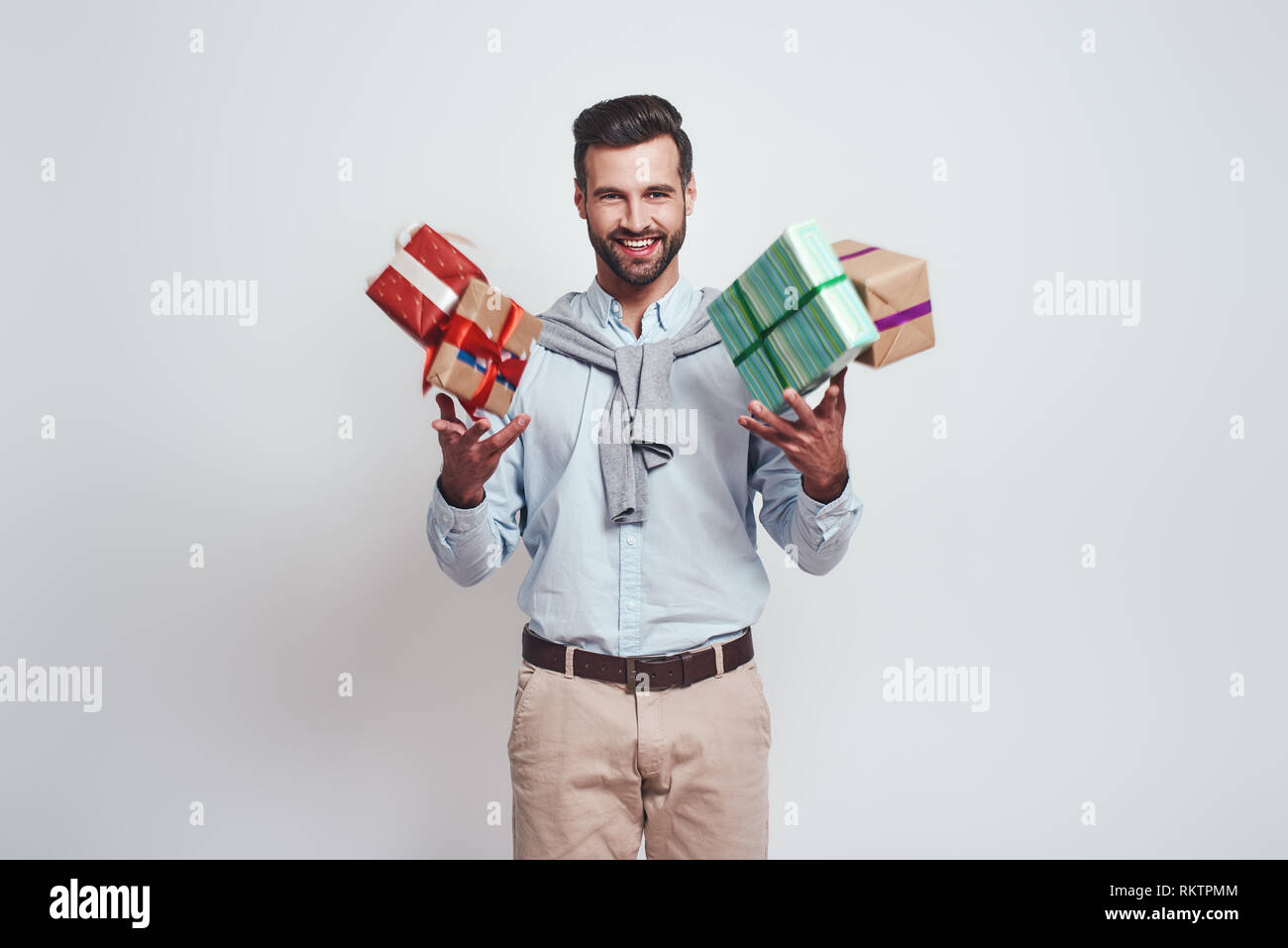 Gifts boom. Cheerful young man is holding gifts and feeling so happy standing on a grey background. Close-up portrait. Preparing for holidays Stock Photo