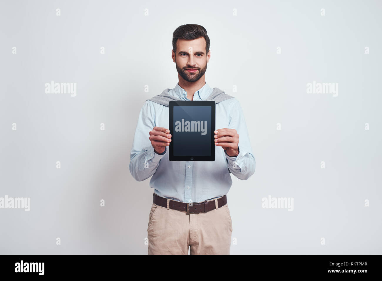 Look over here. An elegant handsome young man holding and showing the screen of a digital touchpad while standing on a grey background. Digital concept. Stock Photo