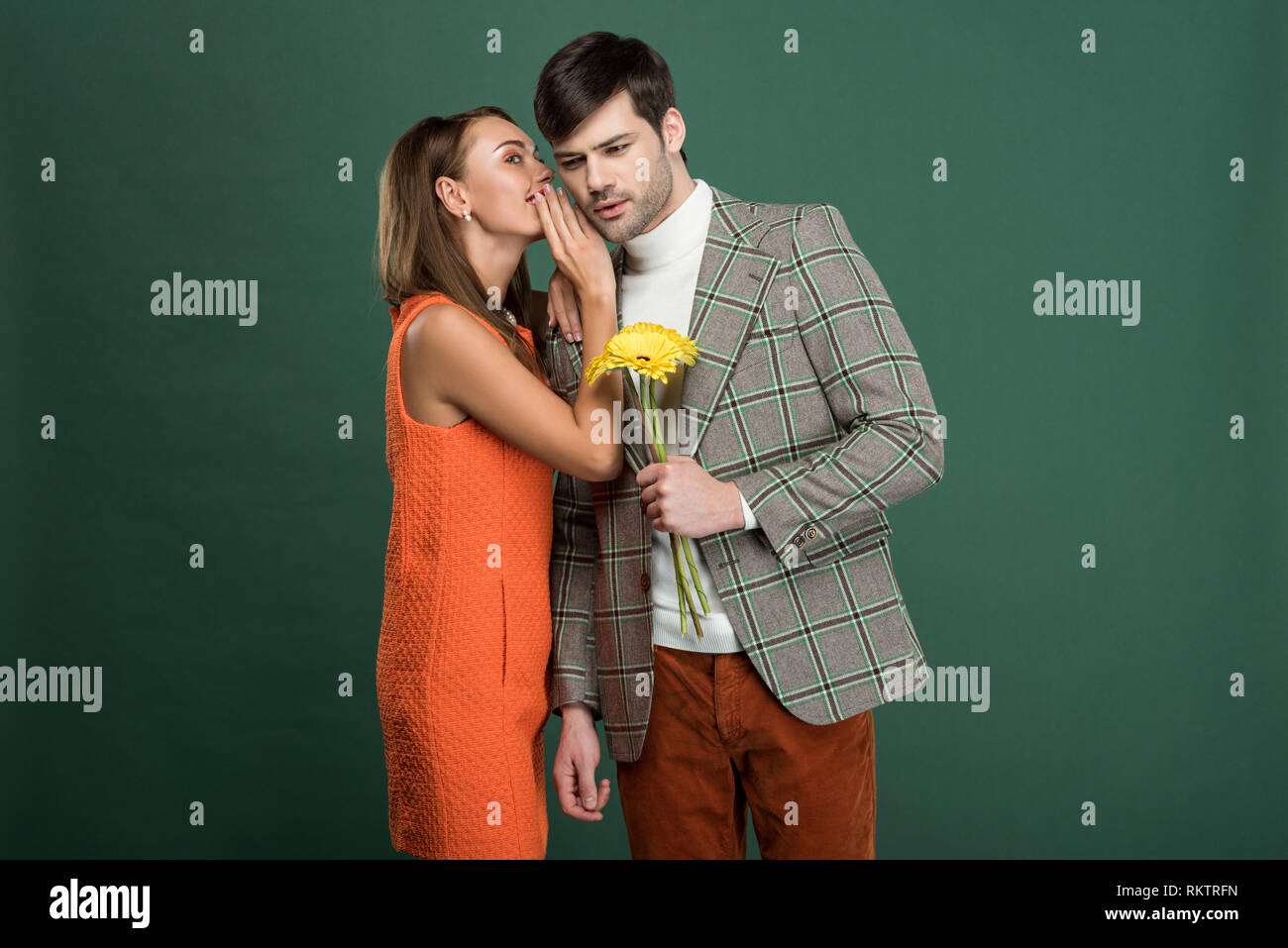 beautiful woman in vintage clothes whispering something to boyfriend with flowers isolated on green Stock Photo