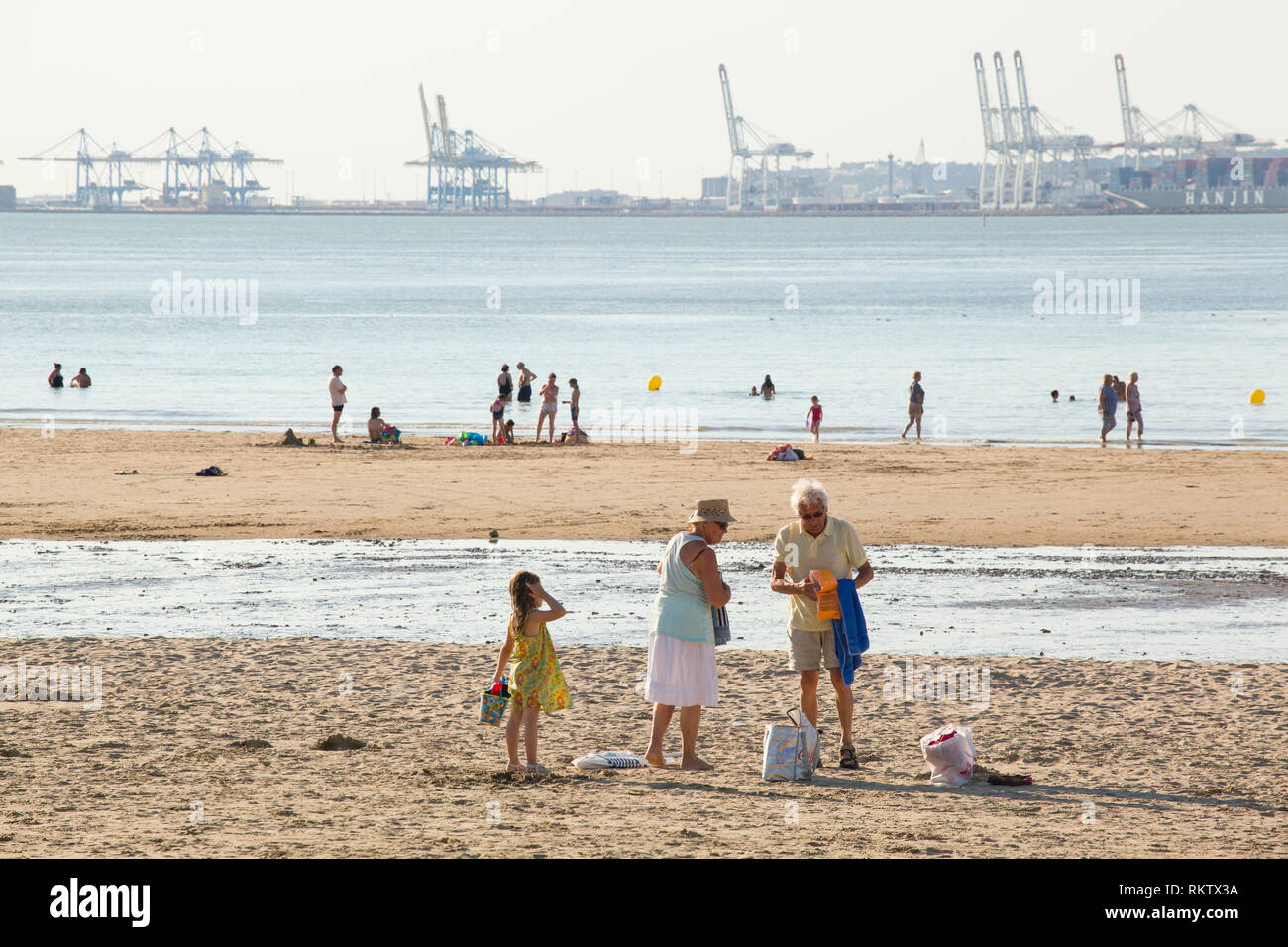 Holidaymakers enjoy the Summer sun on the beach at Plage de Butin, Honfleur, France with the gantry cranes of container port of Le Havre across the Se Stock Photo