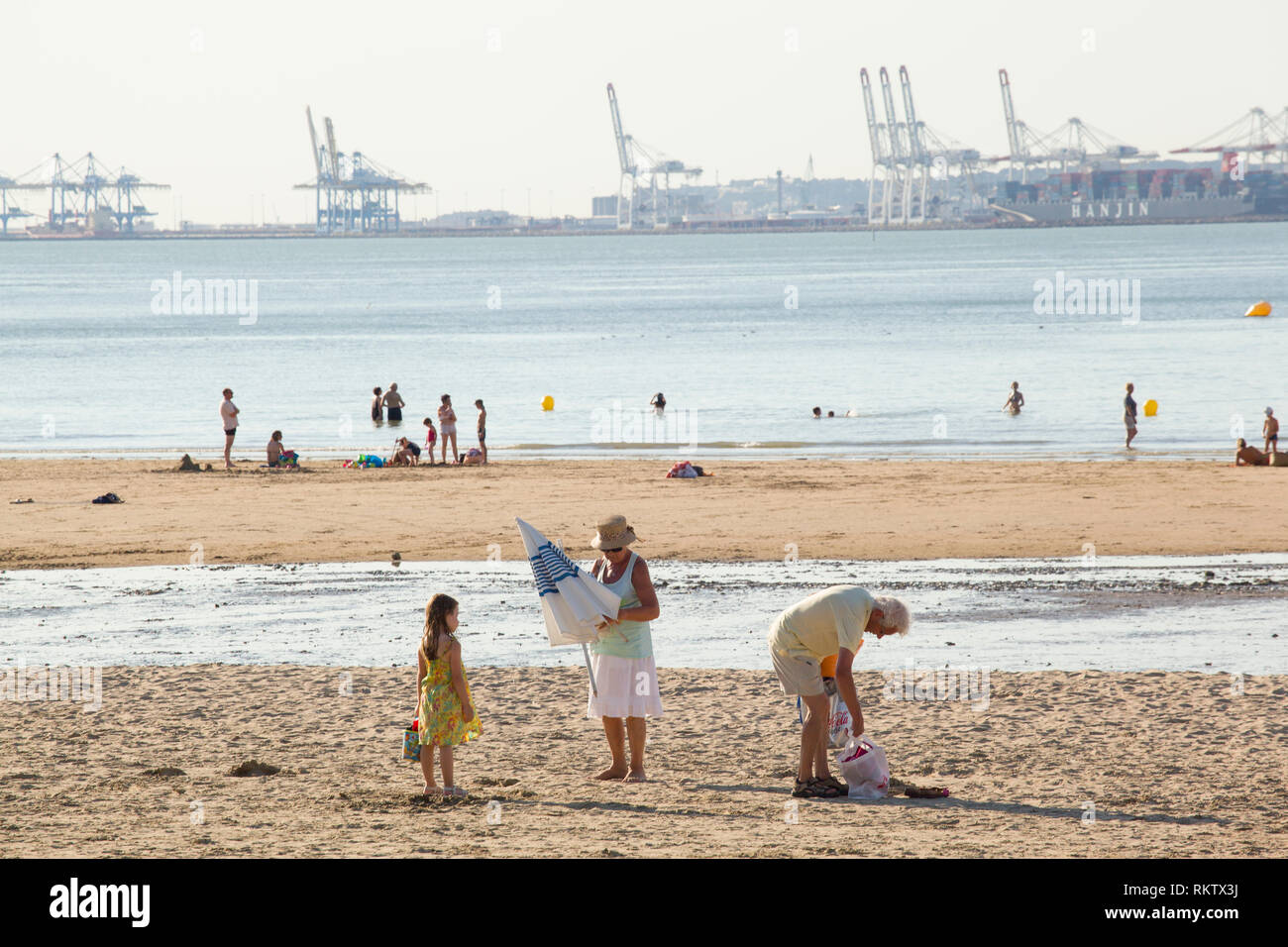 Holidaymakers enjoy the Summer sun on the beach at Plage de Butin, Honfleur, France with the gantry cranes of container port of Le Havre across the Se Stock Photo