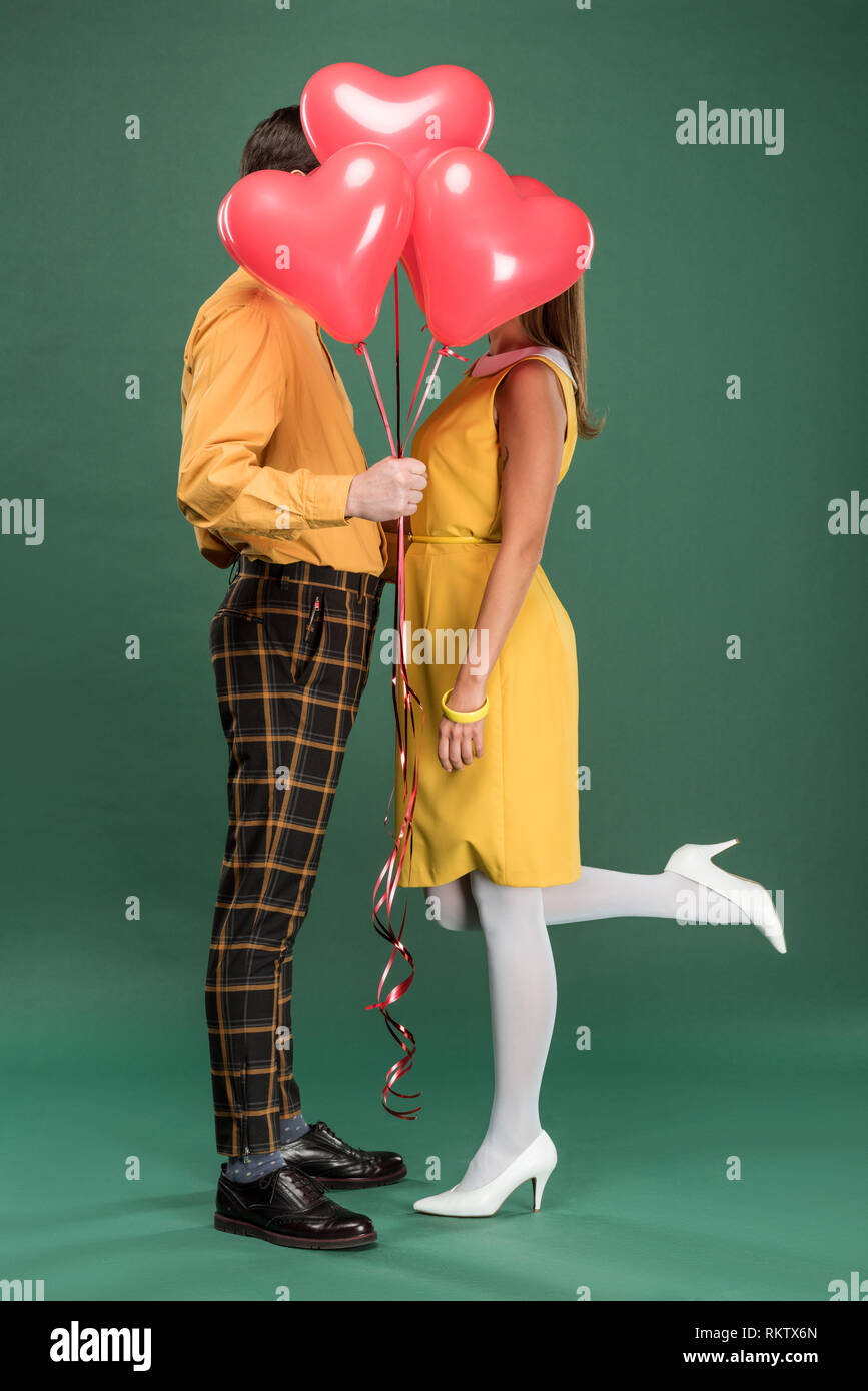 couple covering faces with heart shaped balloons on green background