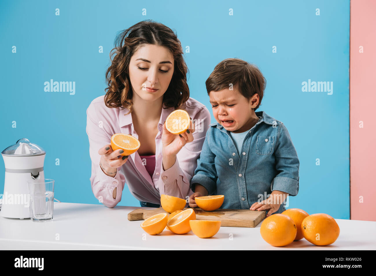 mother holding cut oranges and little son crying while standing at kitchen table on bicolor background Stock Photo