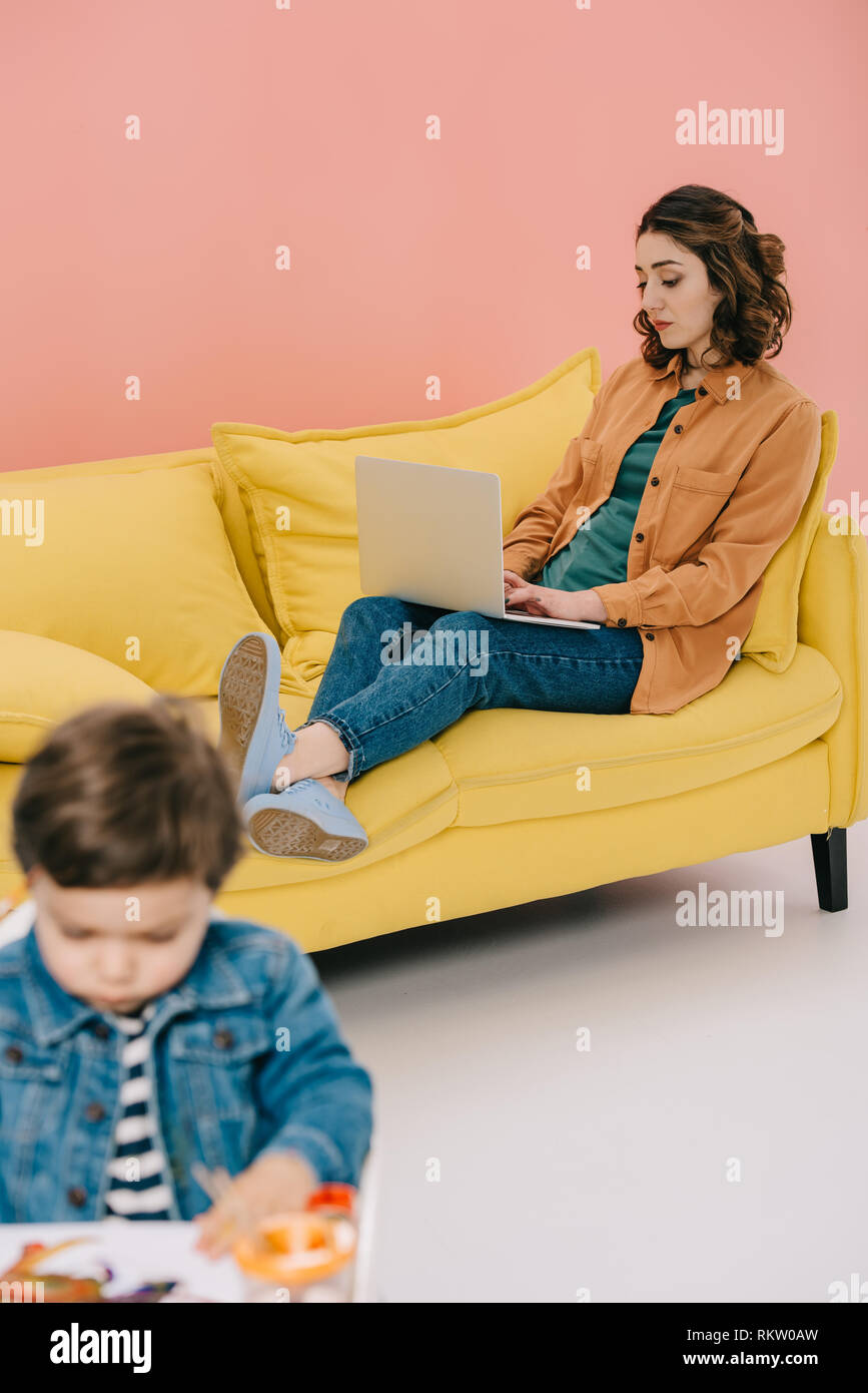 Mother Sitting On Yellow Sofa And Using Laptop While Little Son