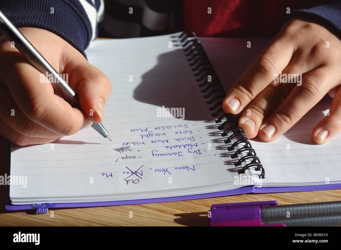 Young woman seated at a desk, updating a list of New Year resolutions in a spiral bound notebook, starting with Dry January. New Year, Old Me Stock Photo