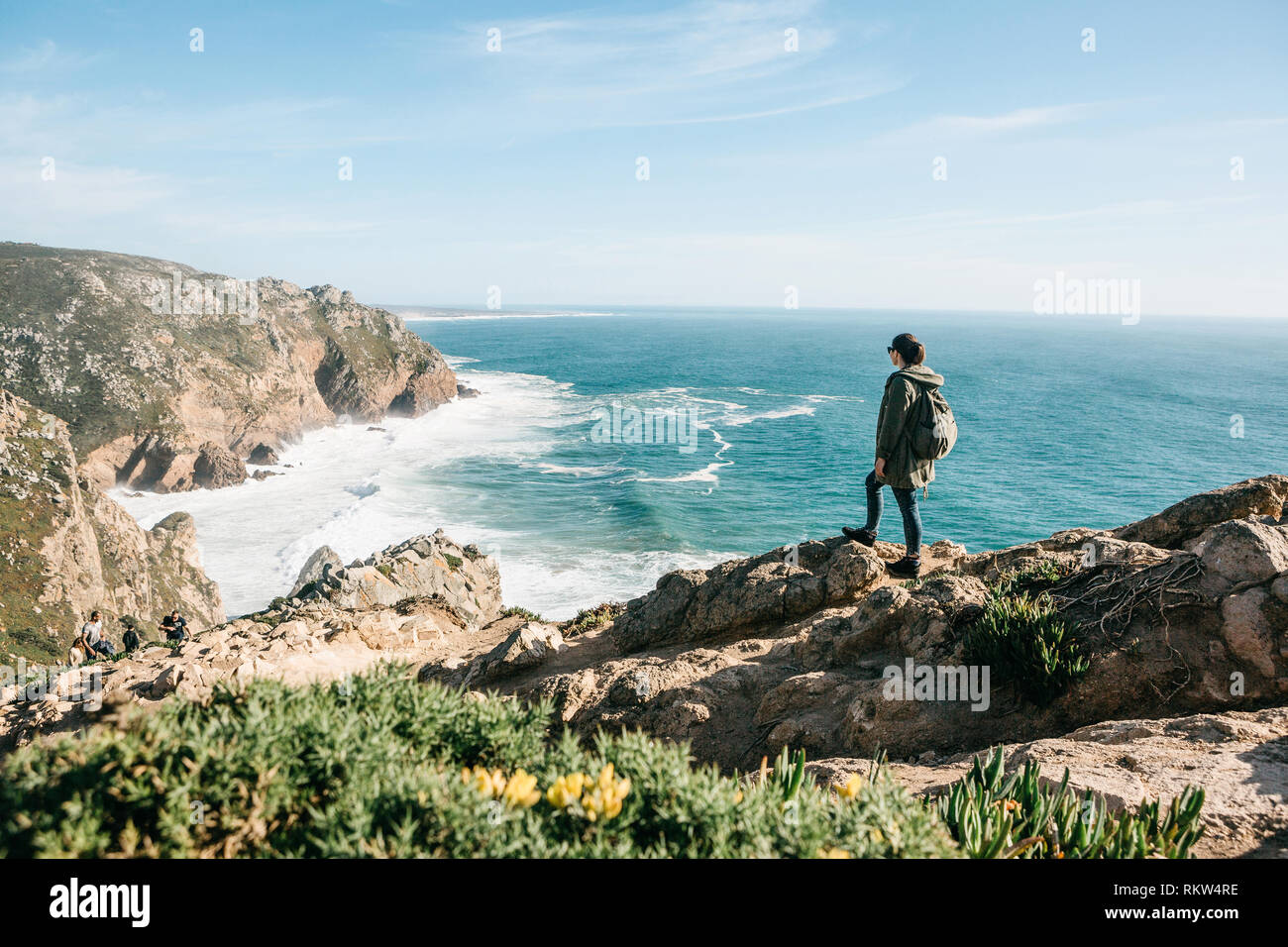 A tourist with a backpack on top of a cliff admires a beautiful view of the Atlantic Ocean in Portugal. Stock Photo