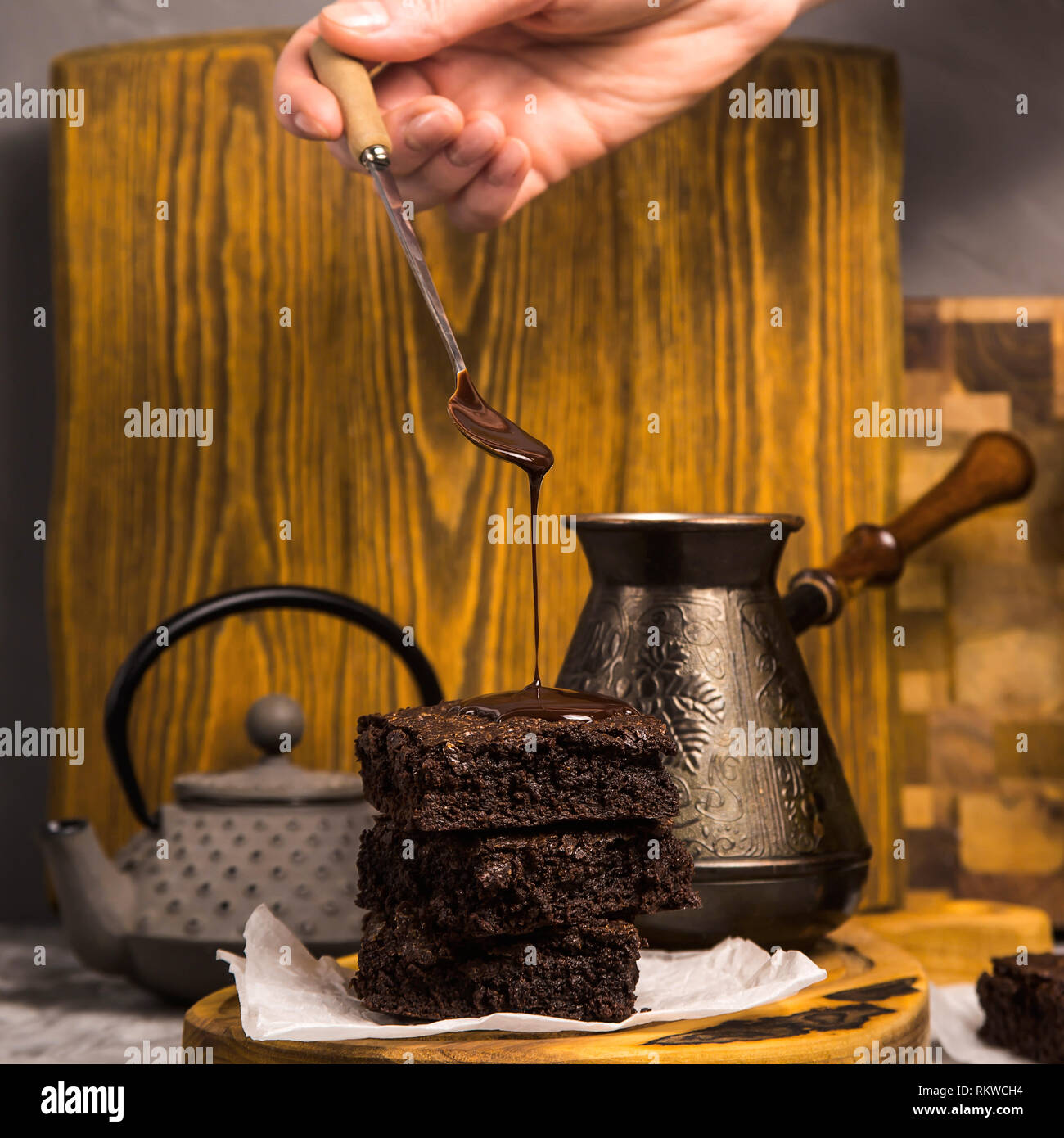 Chocolate is poured from a spoon onto a pile of brownie pieces on a wooden board in a rustic style. Stock Photo