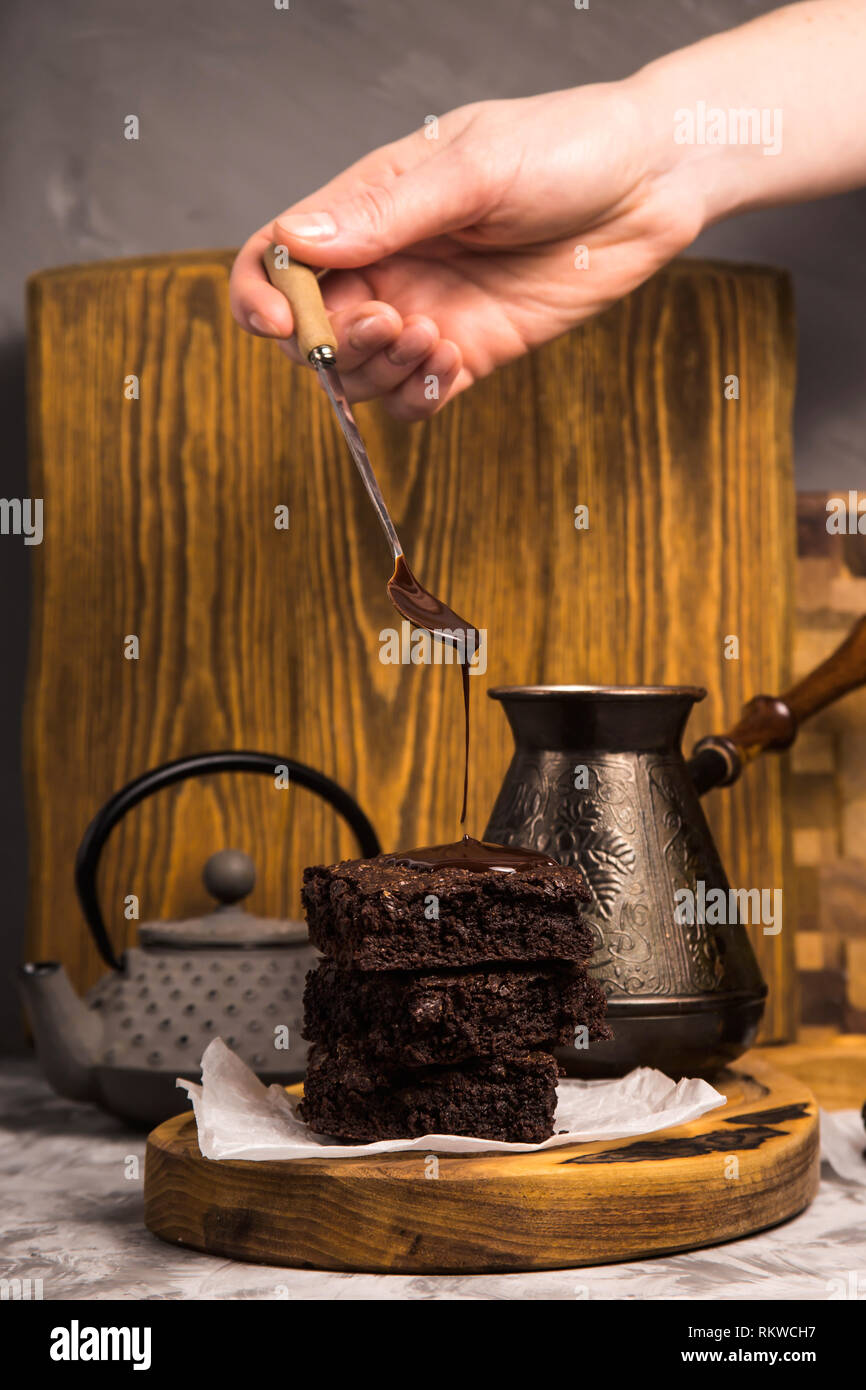 Chocolate is poured from a spoon onto a pile of brownie pieces on a wooden board in a rustic style. Stock Photo