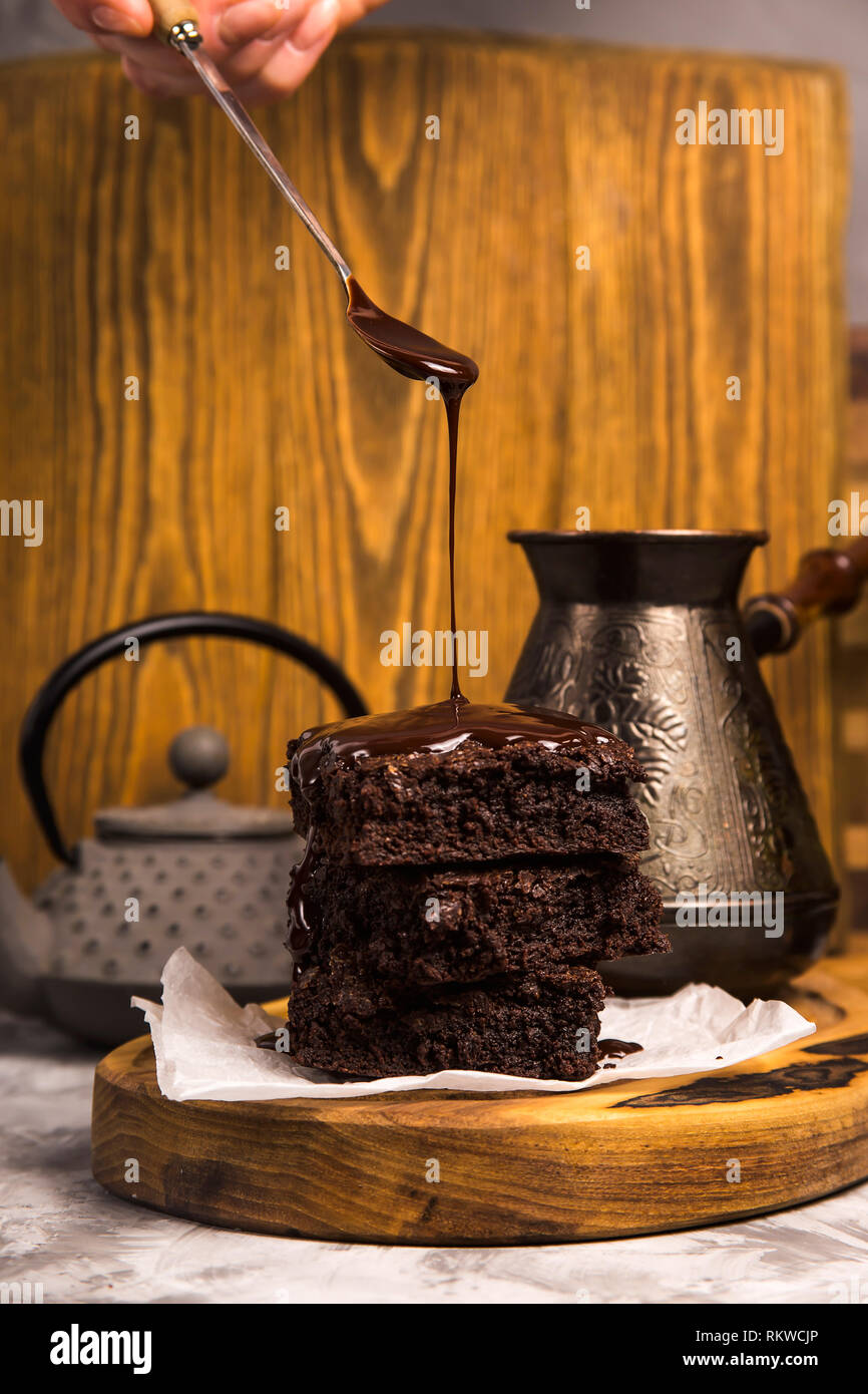 Chocolate is poured from a spoon onto a pile of brownie pieces on a wooden board in a rustic style. Stock Photo
