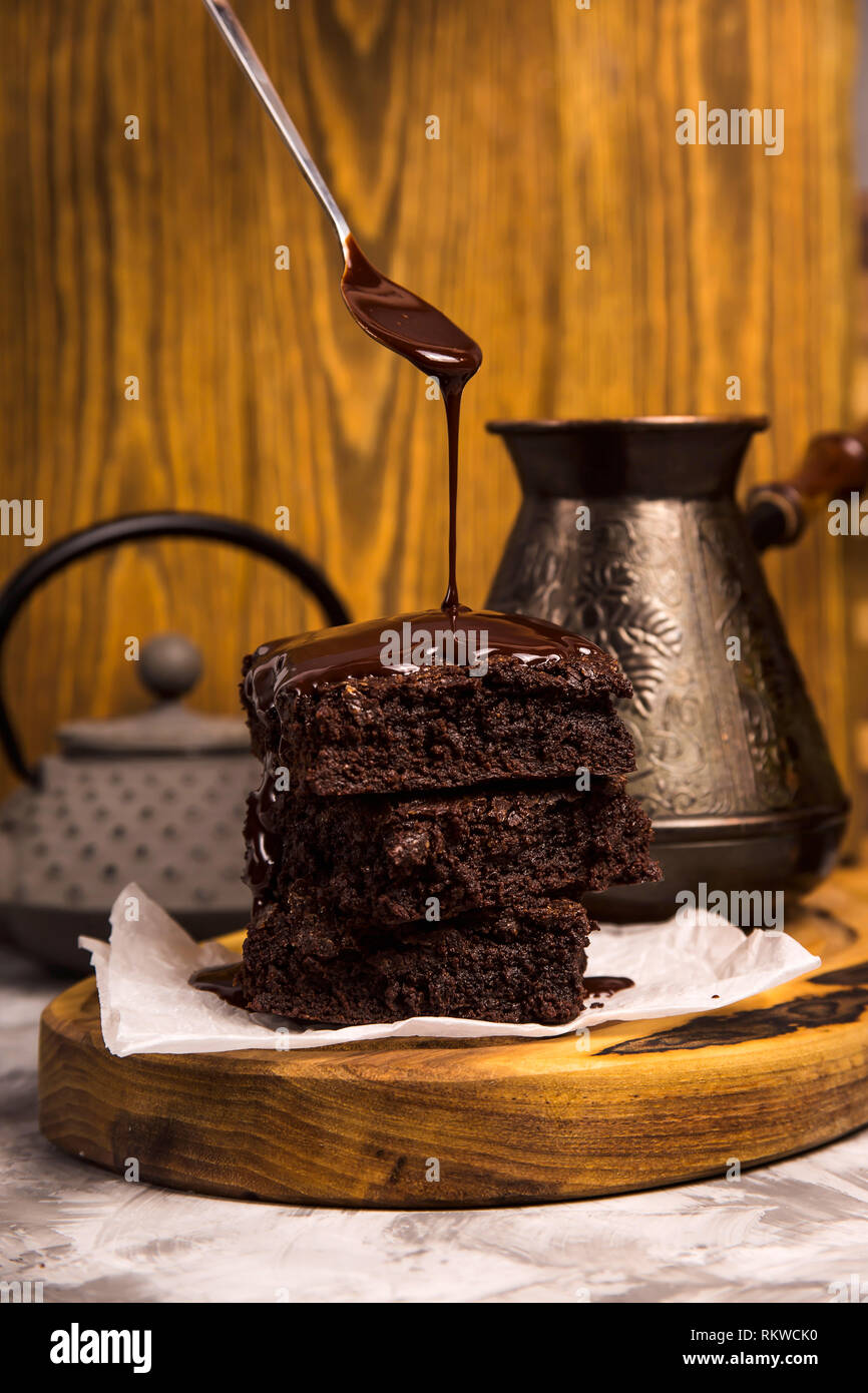 Chocolate is poured from a spoon onto a pile of brownie pieces on a wooden board in a rustic style. Stock Photo