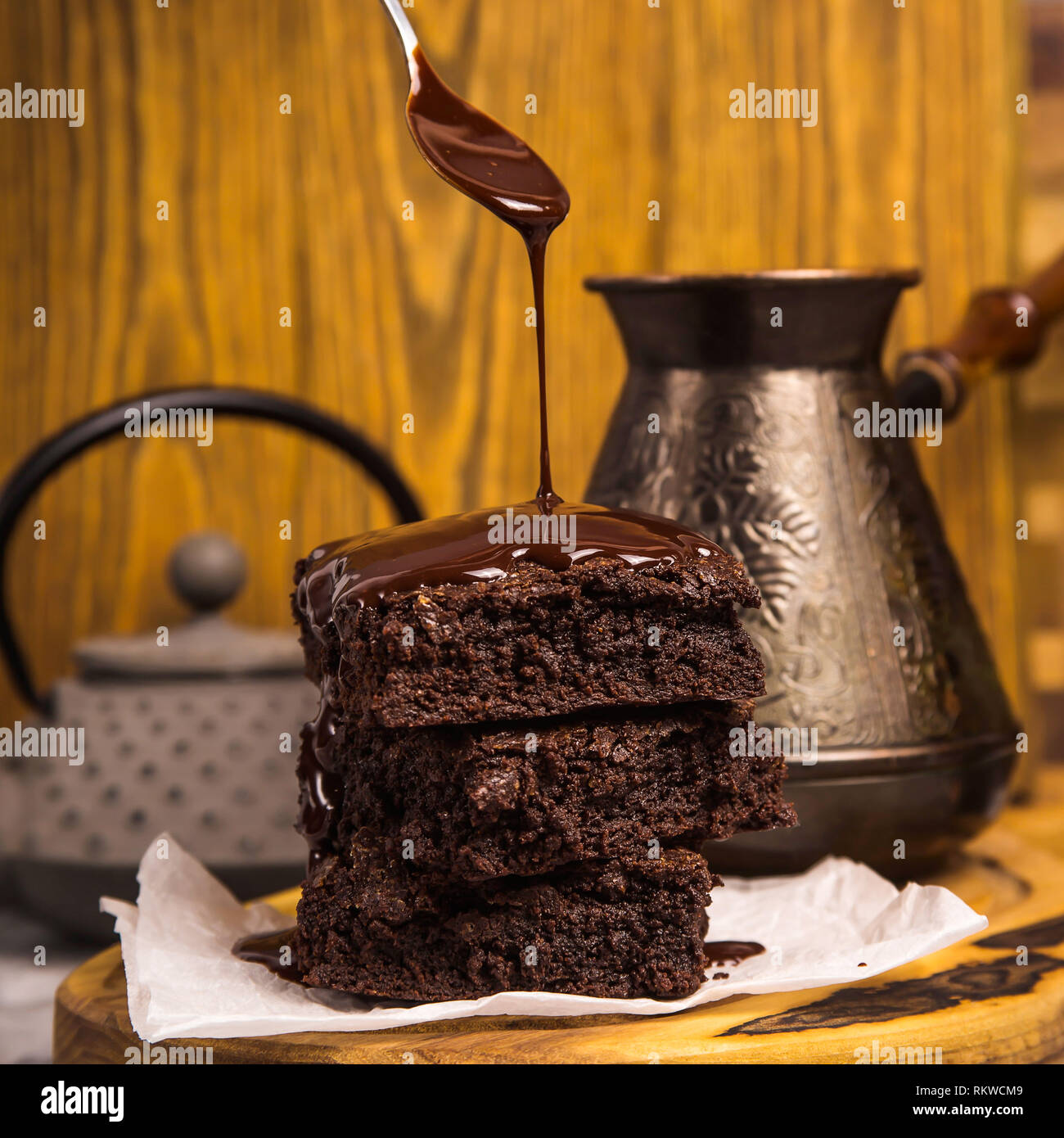 Chocolate is poured from a spoon onto a pile of brownie pieces on a wooden board in a rustic style. Stock Photo