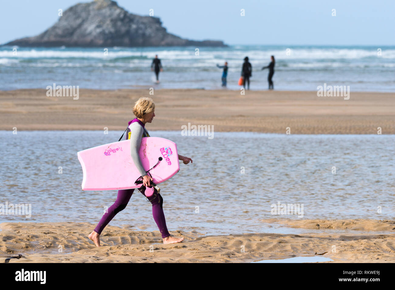 A mature female holidaymaker in a wetsuit carrying a pink body boogie board walking along the shoreline at Crantock Beach in Newquay in Cornwall. Stock Photo